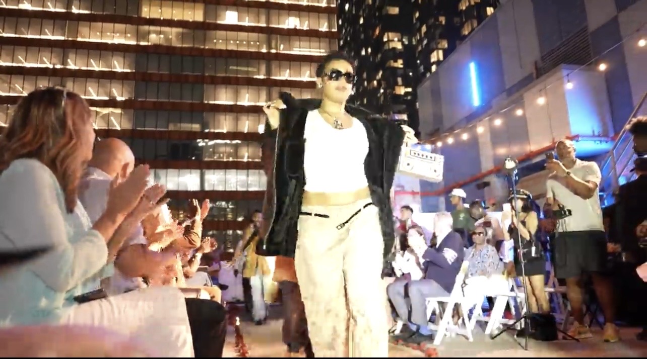 Model walking the Hip Hop Closet rooftop runway in a white tank top and black fur jacket draped over the shoulders, carrying a boombox-style bag.
