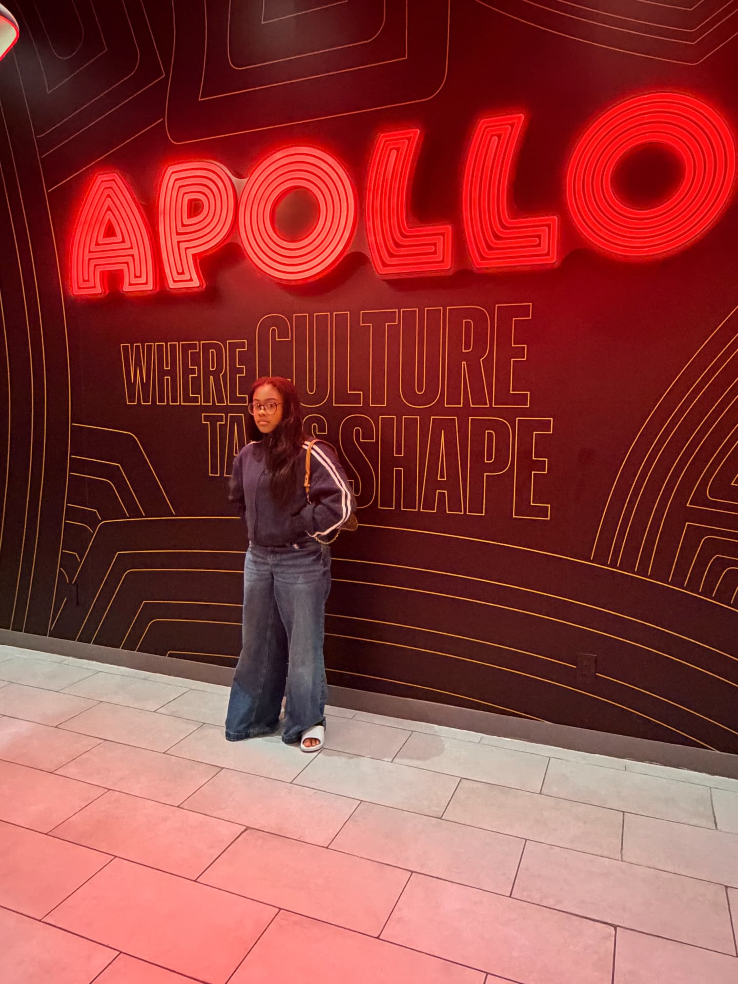 Actress and model Aksana Antonius standing beneath the illuminated red APOLLO — Where Culture Takes Shape sign in Harlem