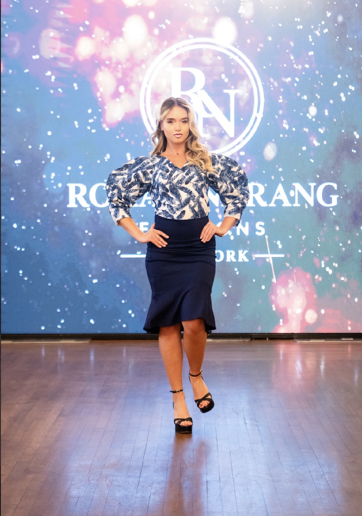 Model in a printed blouse and navy skirt walking the Season 05 runway