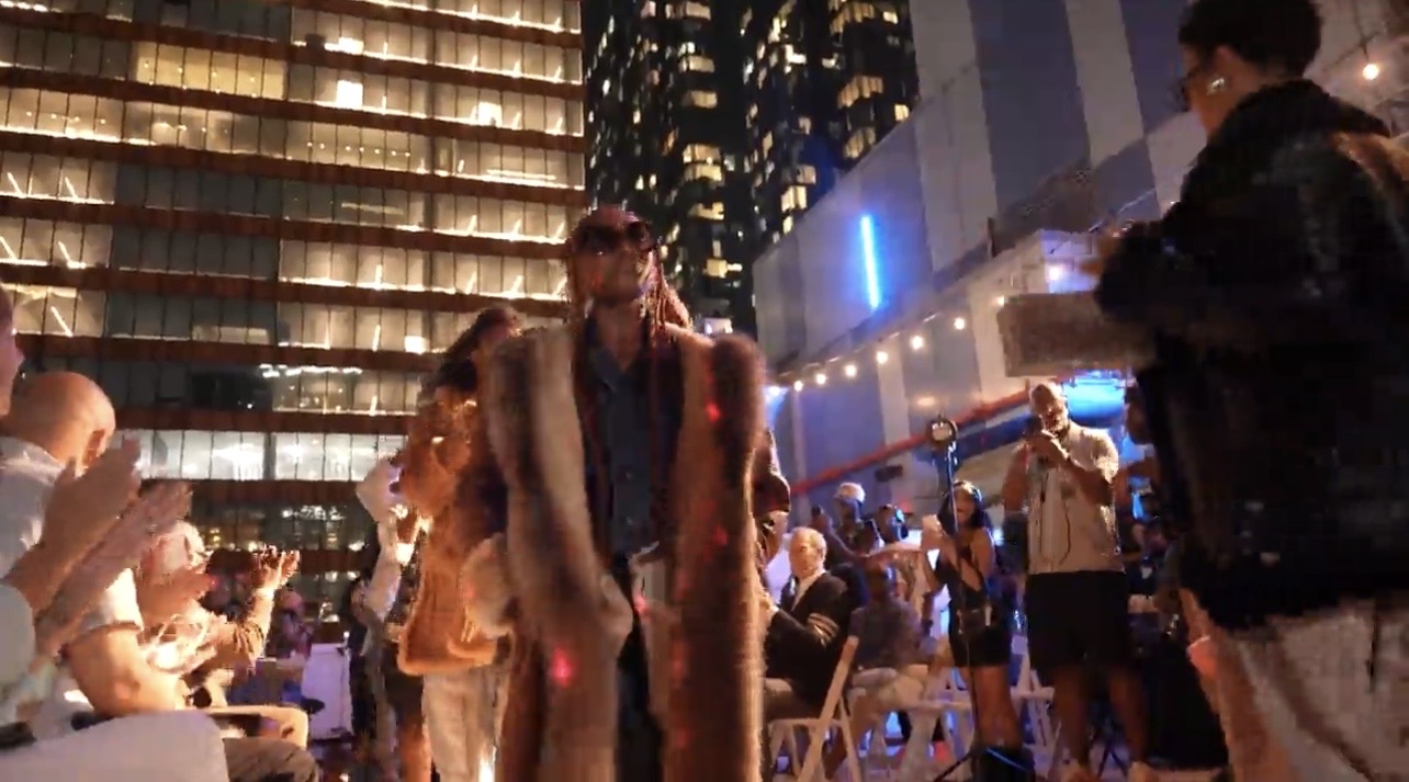 Model walking the Hip Hop Closet rooftop runway at night in a long fur coat and sunglasses, with the New York City skyline behind.