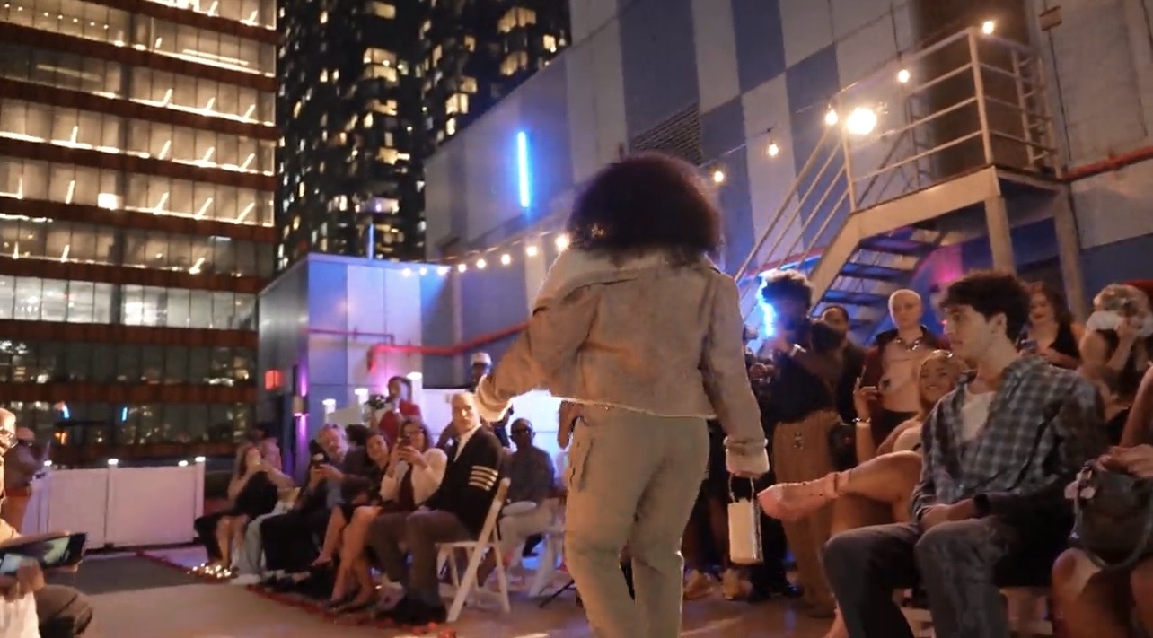 Model on the Hip Hop Closet rooftop runway in a denim jacket and cargo pants, holding a white bag, with the audience on either side.